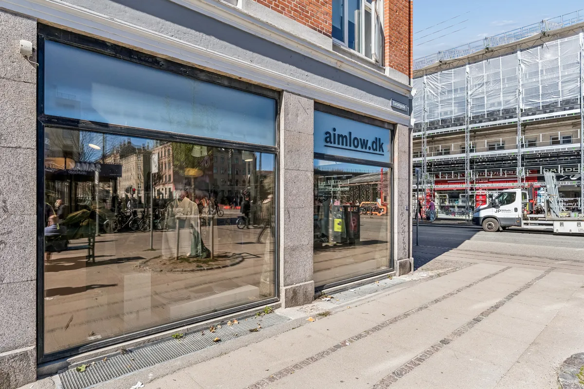 Butiksvinduer med refleksioner af gaden, trafik og mennesker. Køretøj og bygningsværk på arbejde i baggrunden.