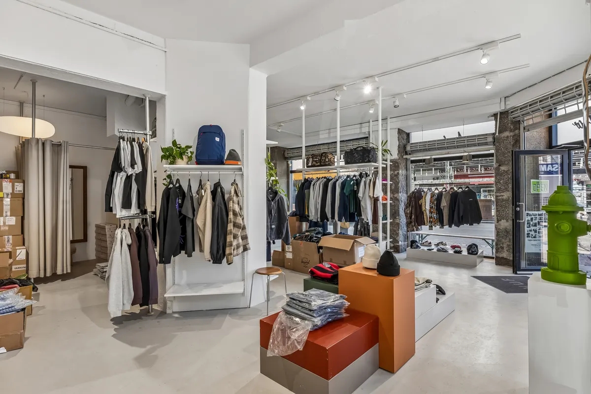 Butik med hængehylder og stolhelv med tøj og rygsække, stol og kuffert på gulvet, stol med hylder, og tøj på reoler og gulvet