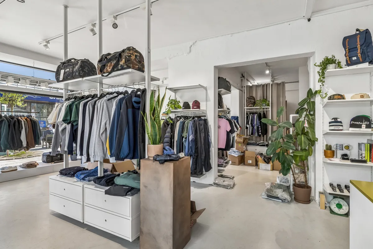 Foto af et moderne butikslokale med hvide vægge, højtloft, hængeklæder og hylder med tøj, tasker og planter, der skaber et hyggeligt og funktionelt indkøbsmiljø