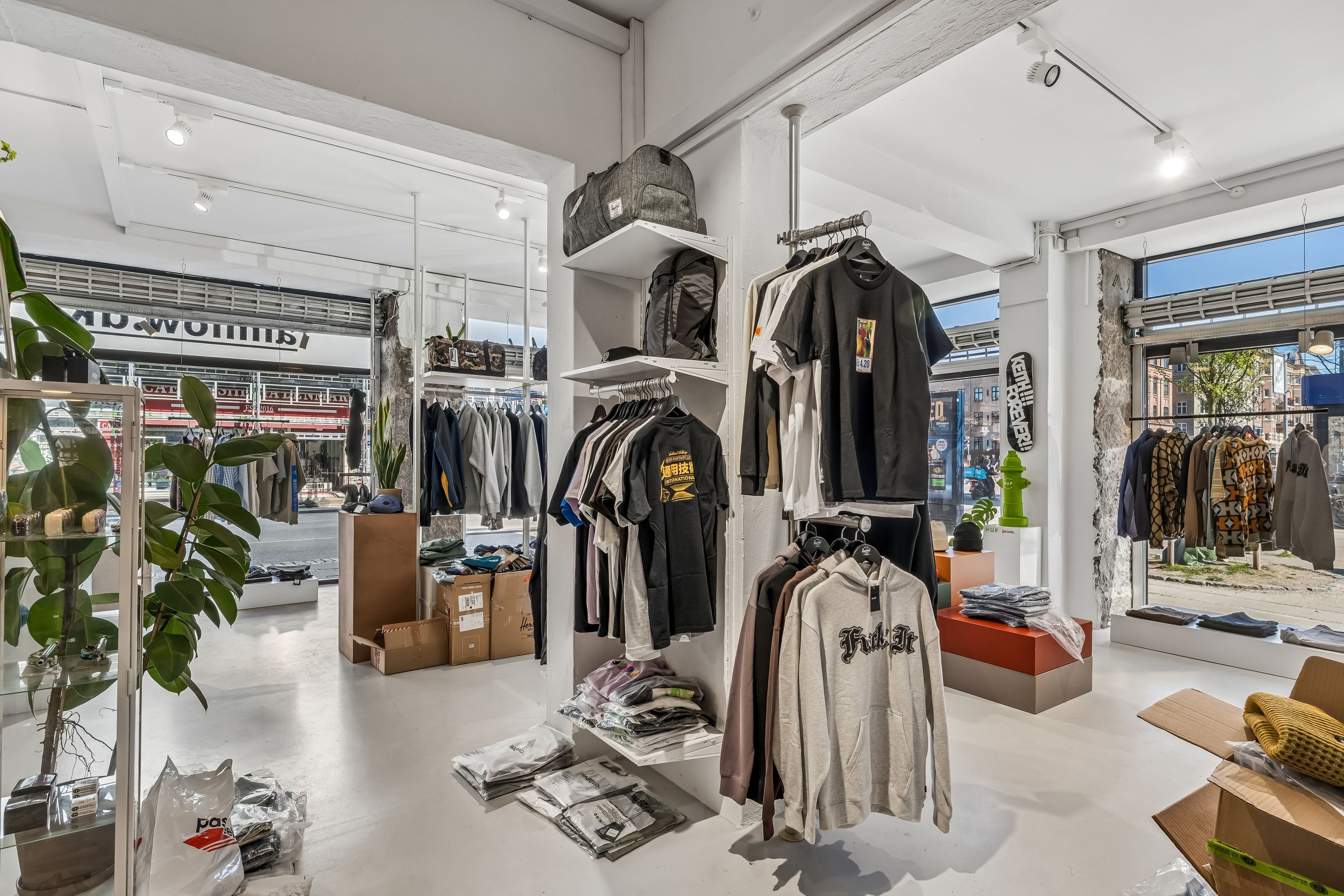 En moderne tøjbutik med tøj på hylder og tøjvægge. Sorte og hvide T-shirts, jeans, sweatshirts og andre tøjvarer. Inde er der også en plante, og der er flere tøjkasses.