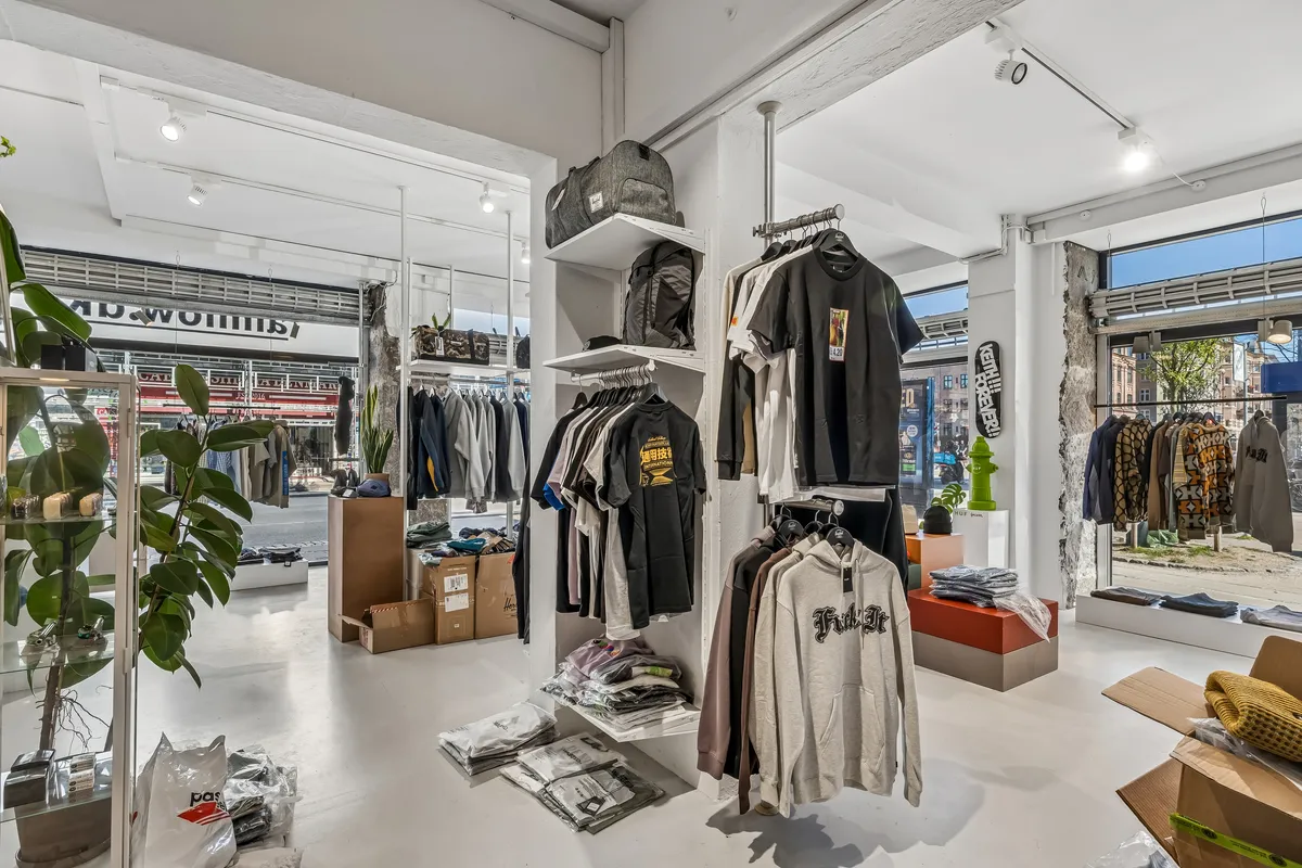 En moderne tøjbutik med tøj på hylder og tøjvægge. Sorte og hvide T-shirts, jeans, sweatshirts og andre tøjvarer. Inde er der også en plante, og der er flere tøjkasses.