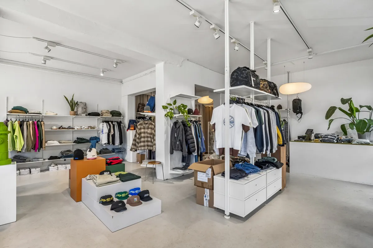 Nymøbleret og veludstyret butik med højt til loftet og moderne indretning. Møbler, klædeskabe, hylder og tekstiler er i hvidt og gråt. Et stort gulvspejl reflekterer rummets indretning