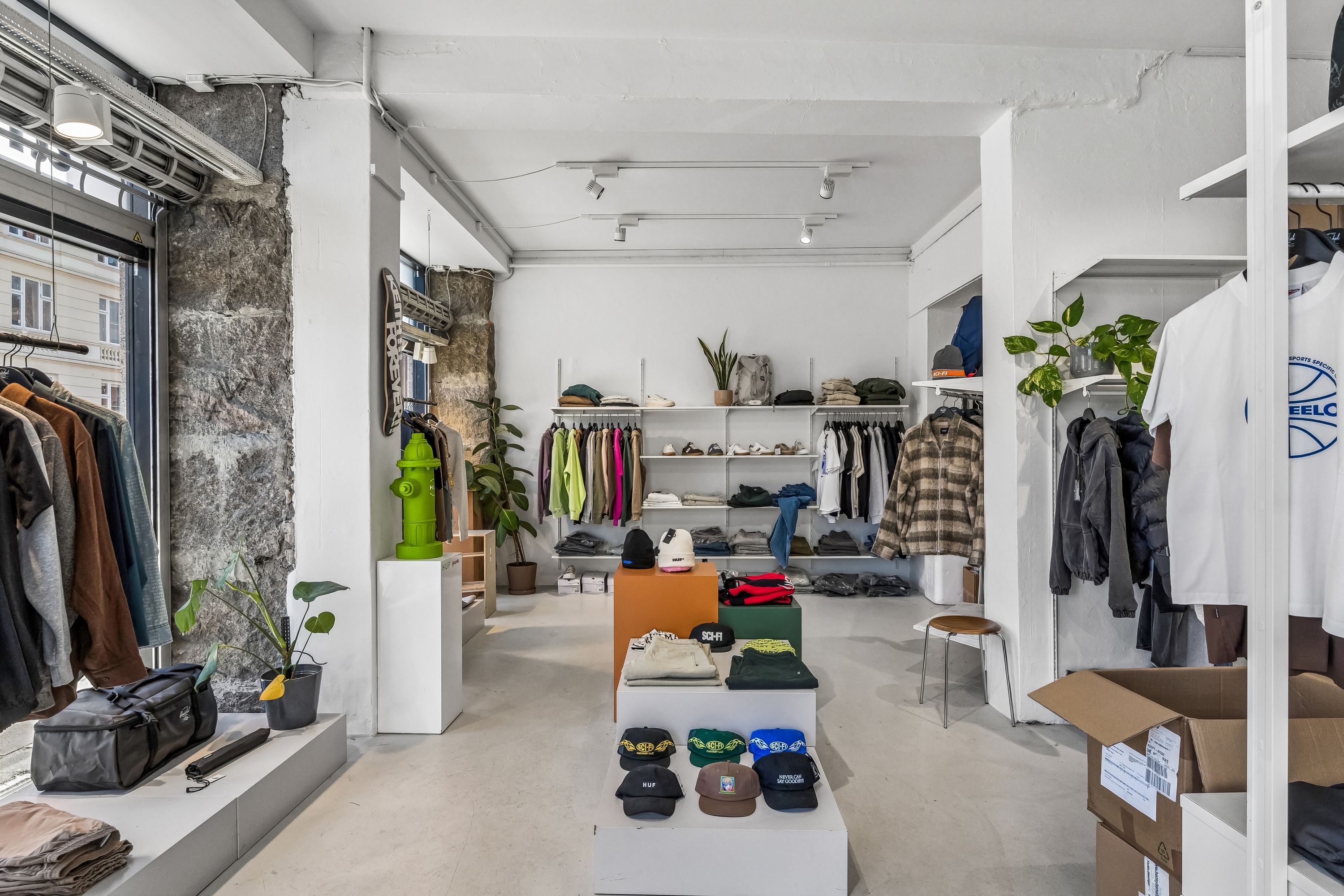 En klædeskabsudstilling med tøj på hylder, hættetrøjer, hatte, sække og planter i en stor og lys butikslokal med lyse gulv og hvide vægge