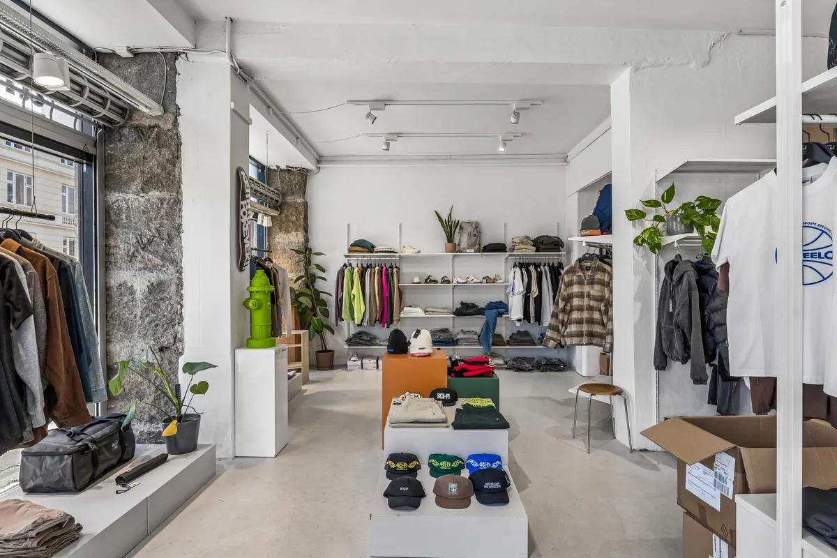 En klædeskabsudstilling med tøj på hylder, hættetrøjer, hatte, sække og planter i en stor og lys butikslokal med lyse gulv og hvide vægge