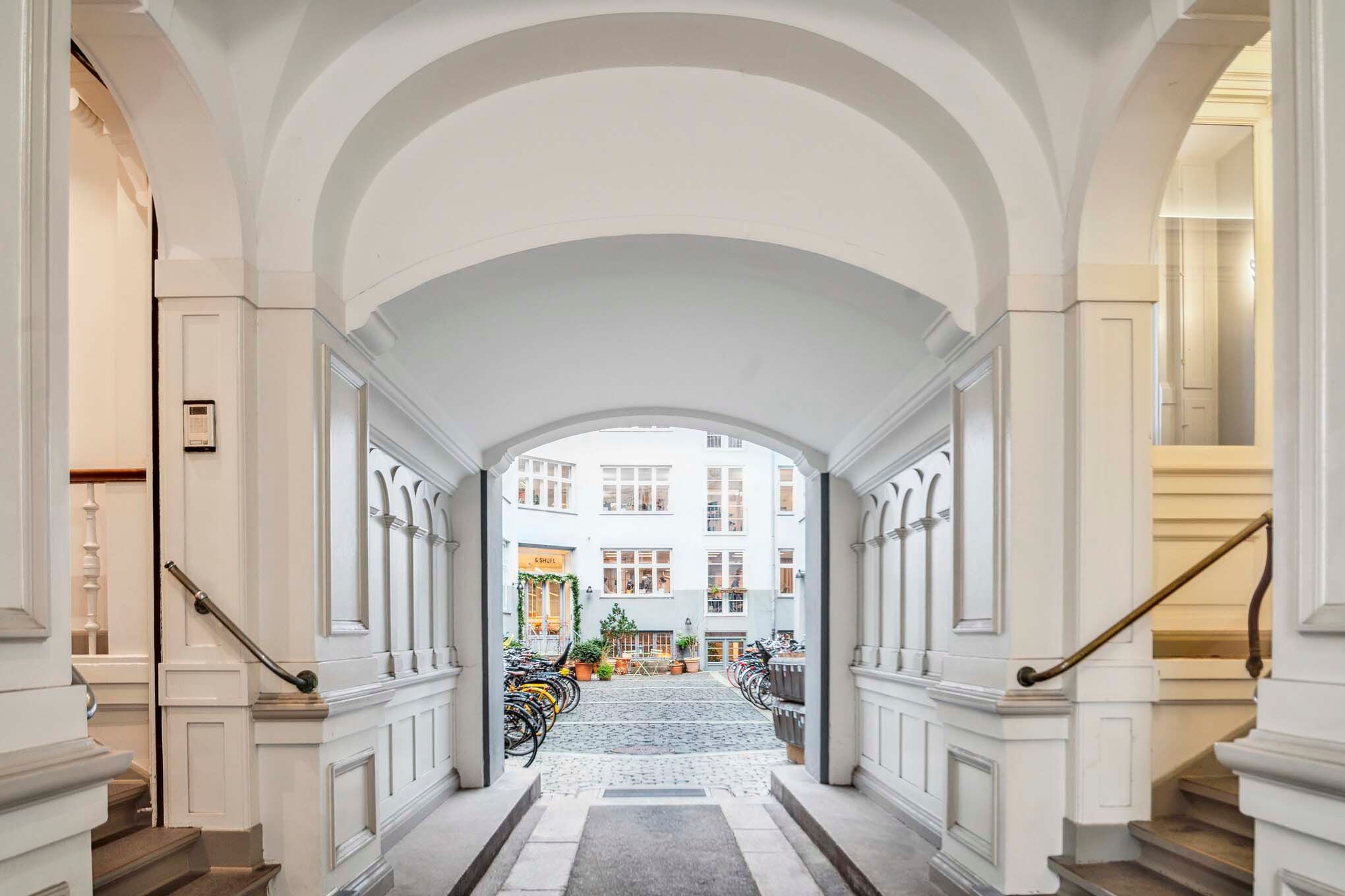 Lys indgangsparti med to trapper og bjælker, indgangsdør og to sidevægge med håndtag og gulvbelægning, samt bagved flere cykler og planter og gulvet er lavet af mursten