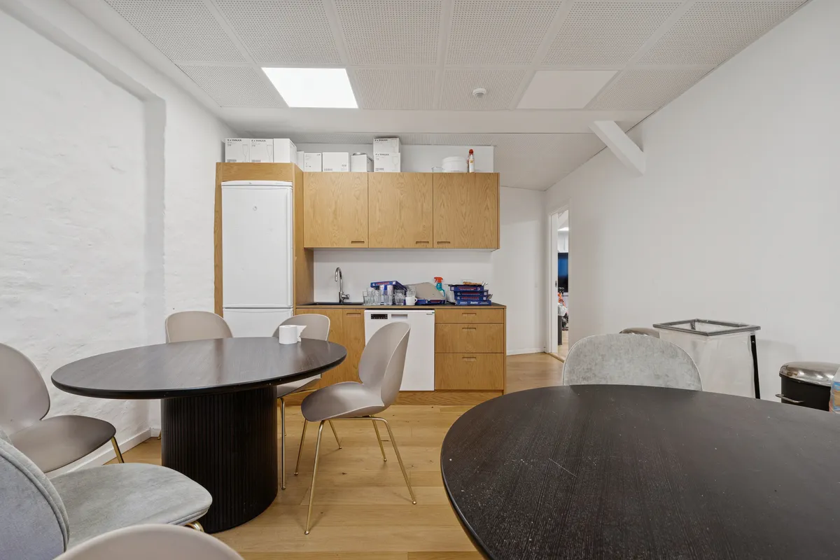 Kontorlejemål med runde borde og høje lyse lofter, køkken med lyse skabe og hvid køleskab og gult gulv
