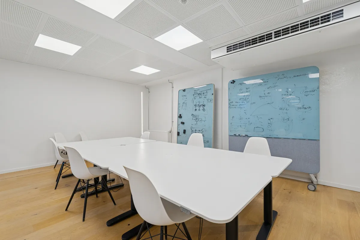 Kontorlejemål med højloftet og lyse loftsvinduer, to store magnetiske whiteboards og moderne møbler i hvidt og sort i et minimalistisk interiør med lyse, trægulve og hvide vægge