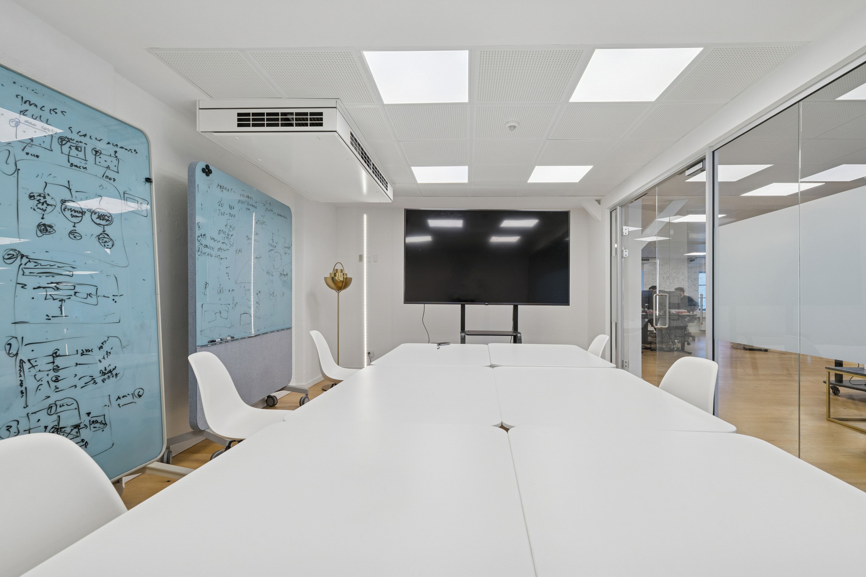 Kontor med hvide vægge, store vinduer, hvide bord og stole samt en stor tv-skærm og hvidt loft med lys