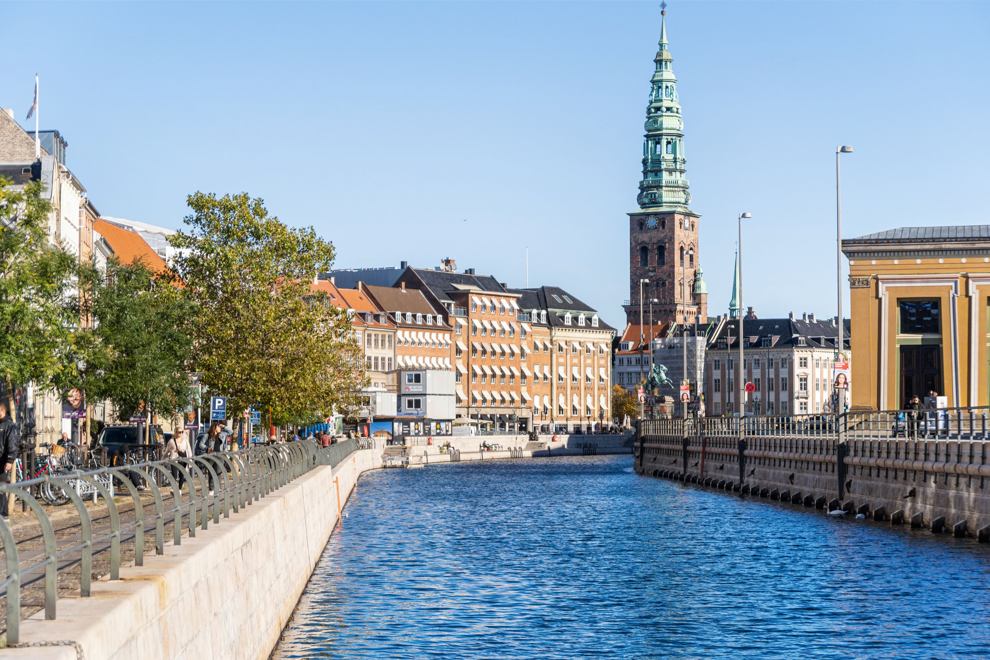 Frederiksholms Kanal med bygninger og statuer, stående mennesker, gågade med cykler, solrigt og tyst vejrum