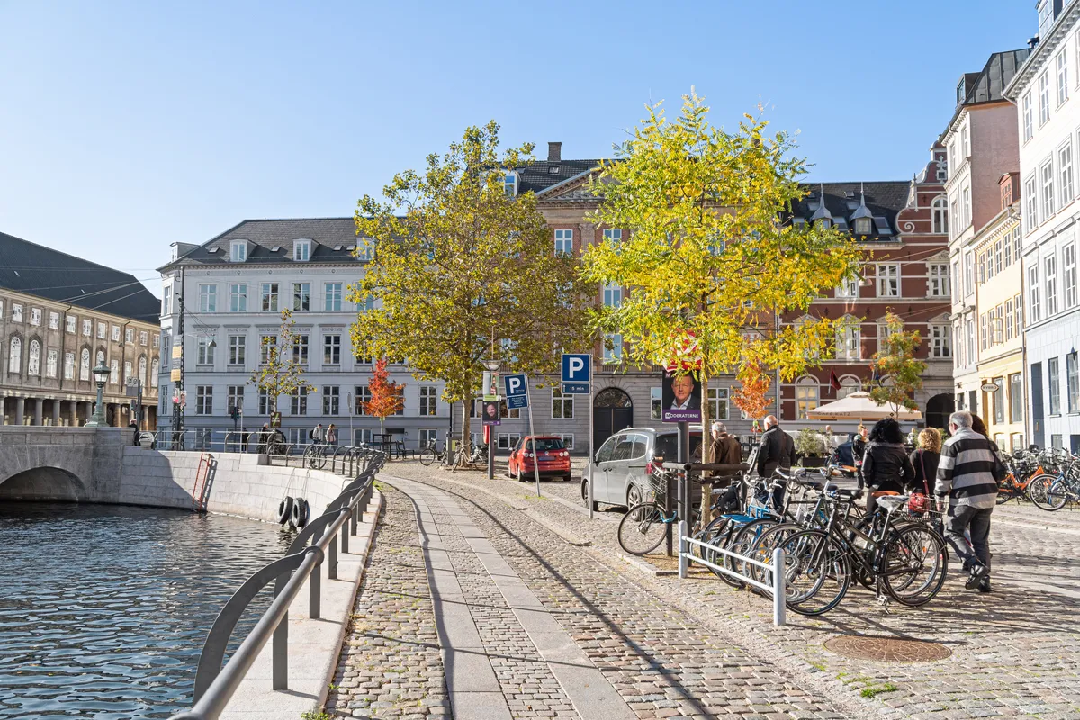 Indre By i København K er præget af bygninger med klassisk arkitektur og grønne træer, mens folk går på broen og parkerer biler langs kanalen