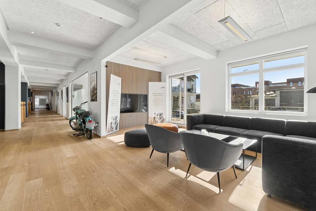 Kontor med industrielt design. Lys stue med sofa, stol og bord. Trægulv og store vinduer. Motorcykel og plakater.