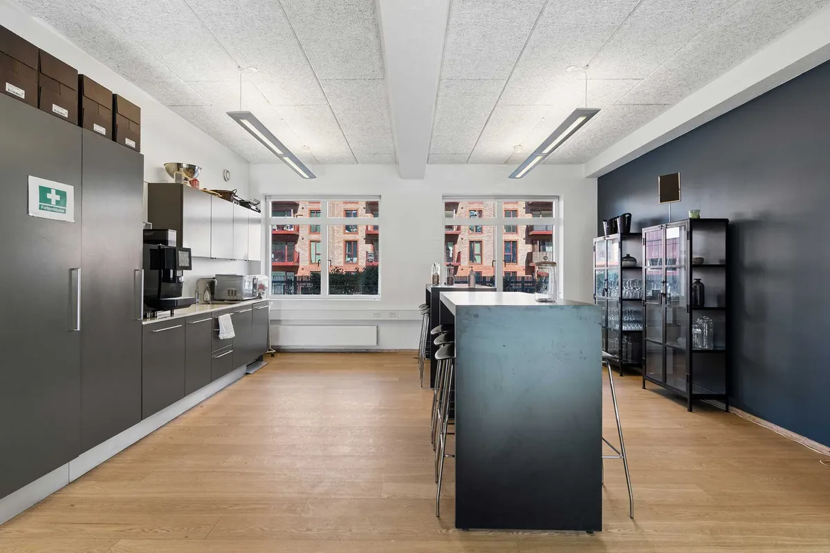 Lyst kontorlejemål med lyse lofter, glasvinduer og trægulv. Væggene er malet i lyse farver og der er en stor glasdør mod et skur.