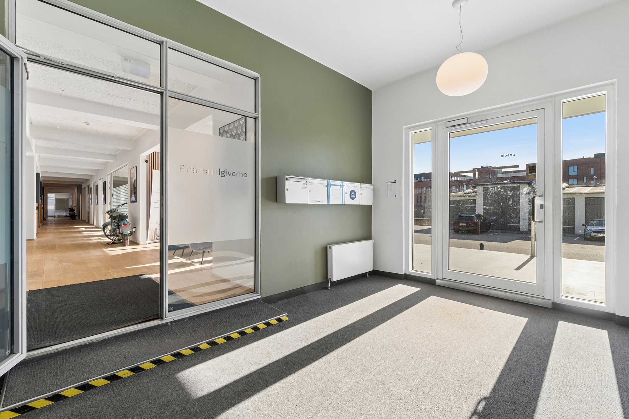 Moderne kontorlejemål med lyse indvendige vægge og store glasruder og lyse refleksioner på gulvet og væggen. Adgangsdør og skorstensrør i baggrunden