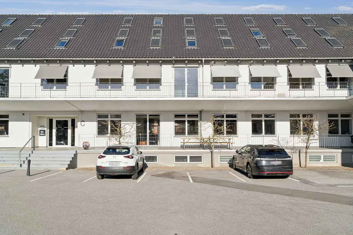 Moderne kontorblok med højt loft og solcellepaneler, to biler parkeret, terrasse med møbelgruppe og træer i forgrunden, skrift C på facaden