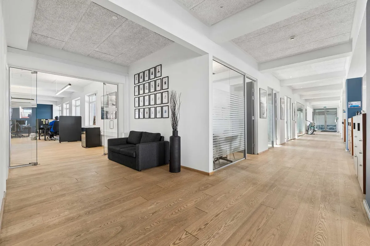 Lys kontorfællesskab med højloftede lokaler og moderne møbler. Indeholder et sovehjørne med lædersofa og trægulv.