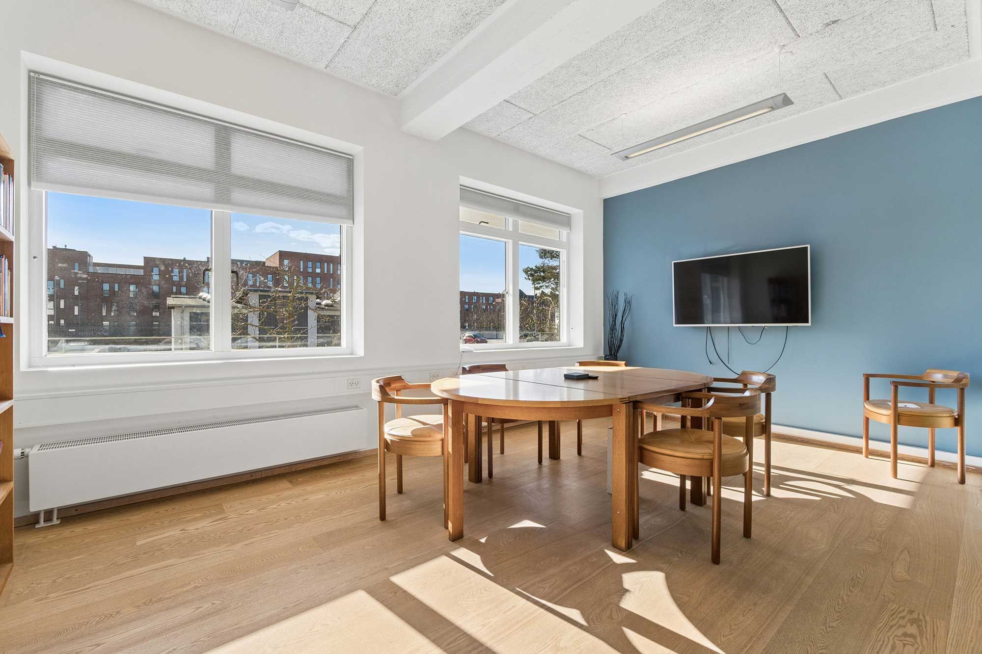 En kontormøbleret med runde bord, stole og TV. Vinduet har gardiner og udsigt til bygninger.