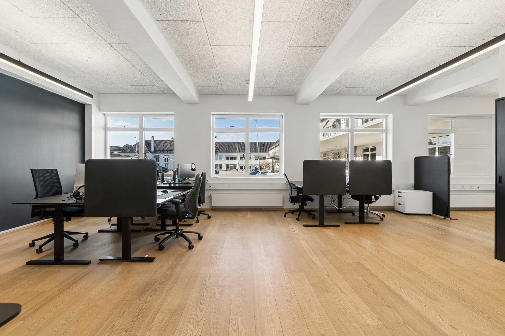 Kontorlokaler med lyse vinduer og moderne møbler og gulvbelægning. Kontorer med moderne skrivebord og stol. Vægge med mørke farver og hvide loftsbjælker.