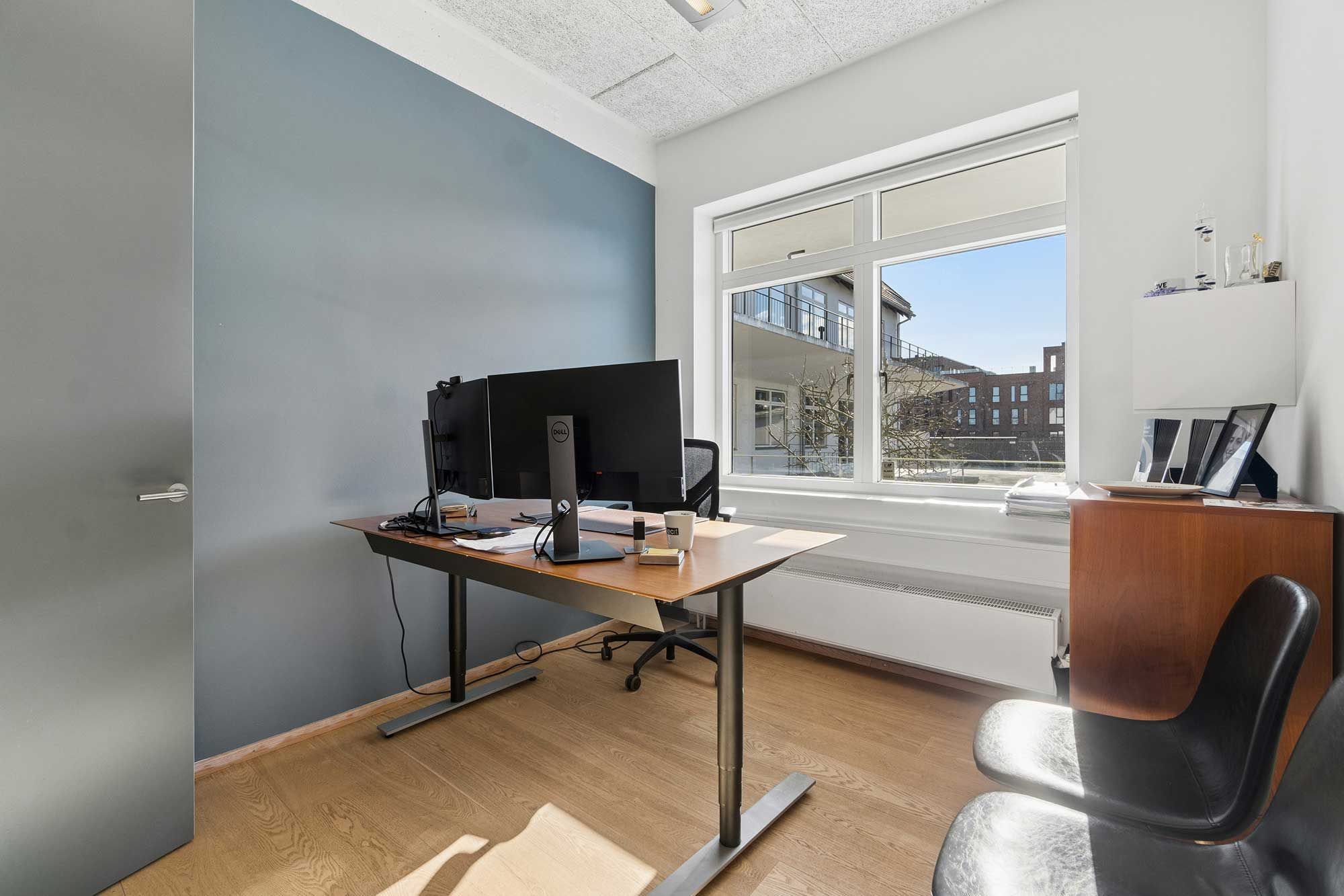 Kontorrum med skrivebord og stole samt monitorer og lyse vinduer med udsigt til bygninger og gader