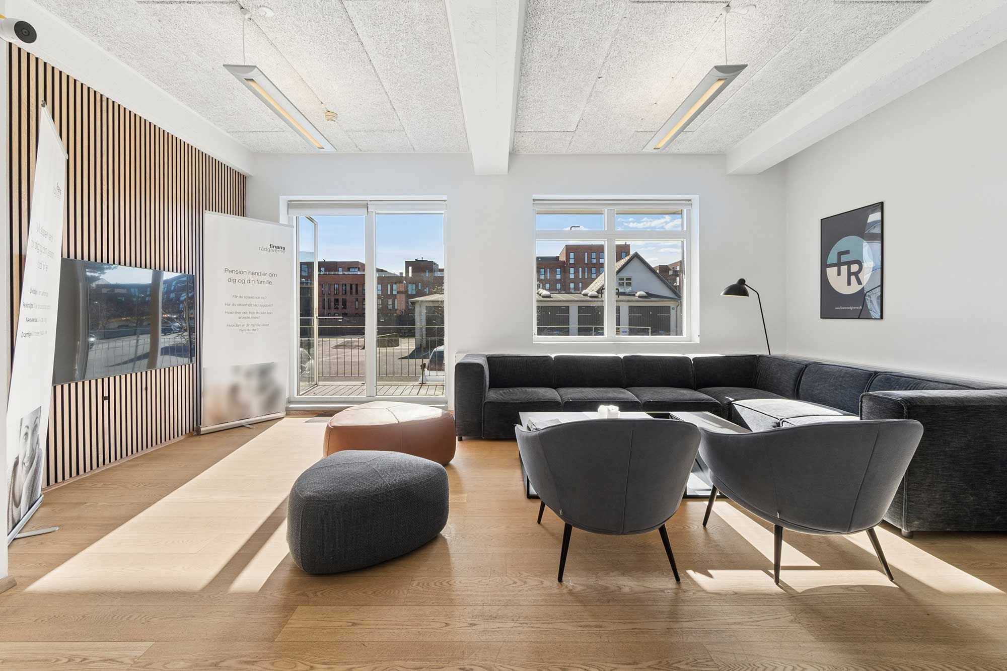 Kontor med L-form sofa, runde læsestol, gulvtæppe, bord, lys trægulv, højt loft, glasdøre, slank lampestol, tavle og tv-skærm på væggen