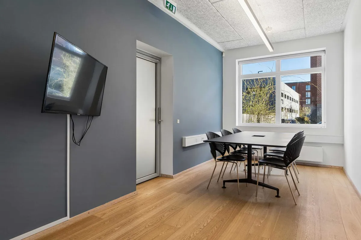 Kontorrum med lyse loftslamper og hvide vægge, trægulv, sort bord og stole, samt en TV-skærm hængende på væggen. Der er også en dør med glas og en vinduesåbning.