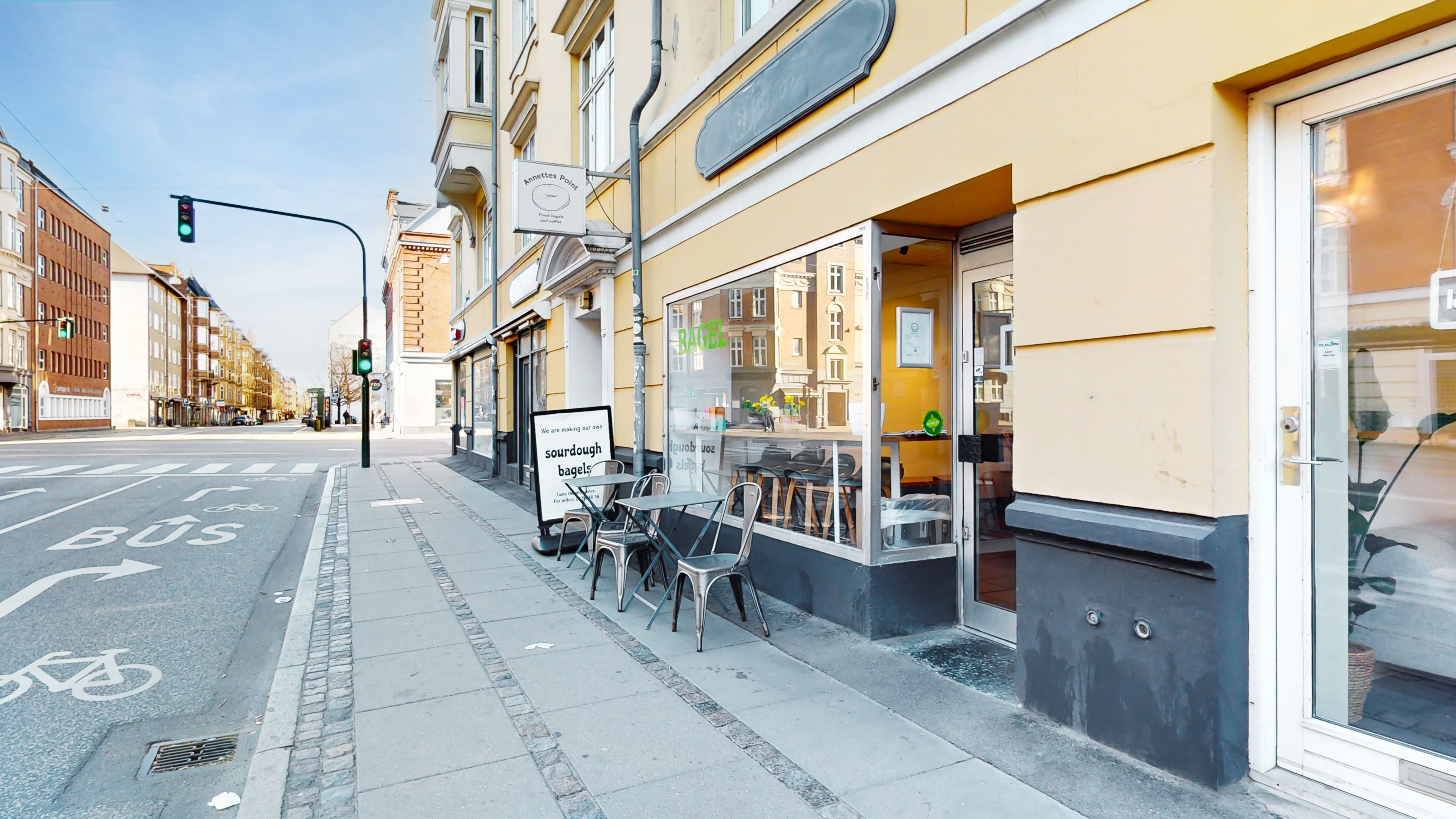 Erhvervslokaler med terrasse og indretning til café eller detailforretning. Træborde og stole med udsigt til gaden og en annonce for surdej bagerier. Lys og rummelige lokaler med store ruder