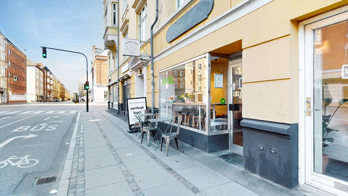 Erhvervslokaler med terrasse og indretning til café eller detailforretning. Træborde og stole med udsigt til gaden og en annonce for surdej bagerier. Lys og rummelige lokaler med store ruder