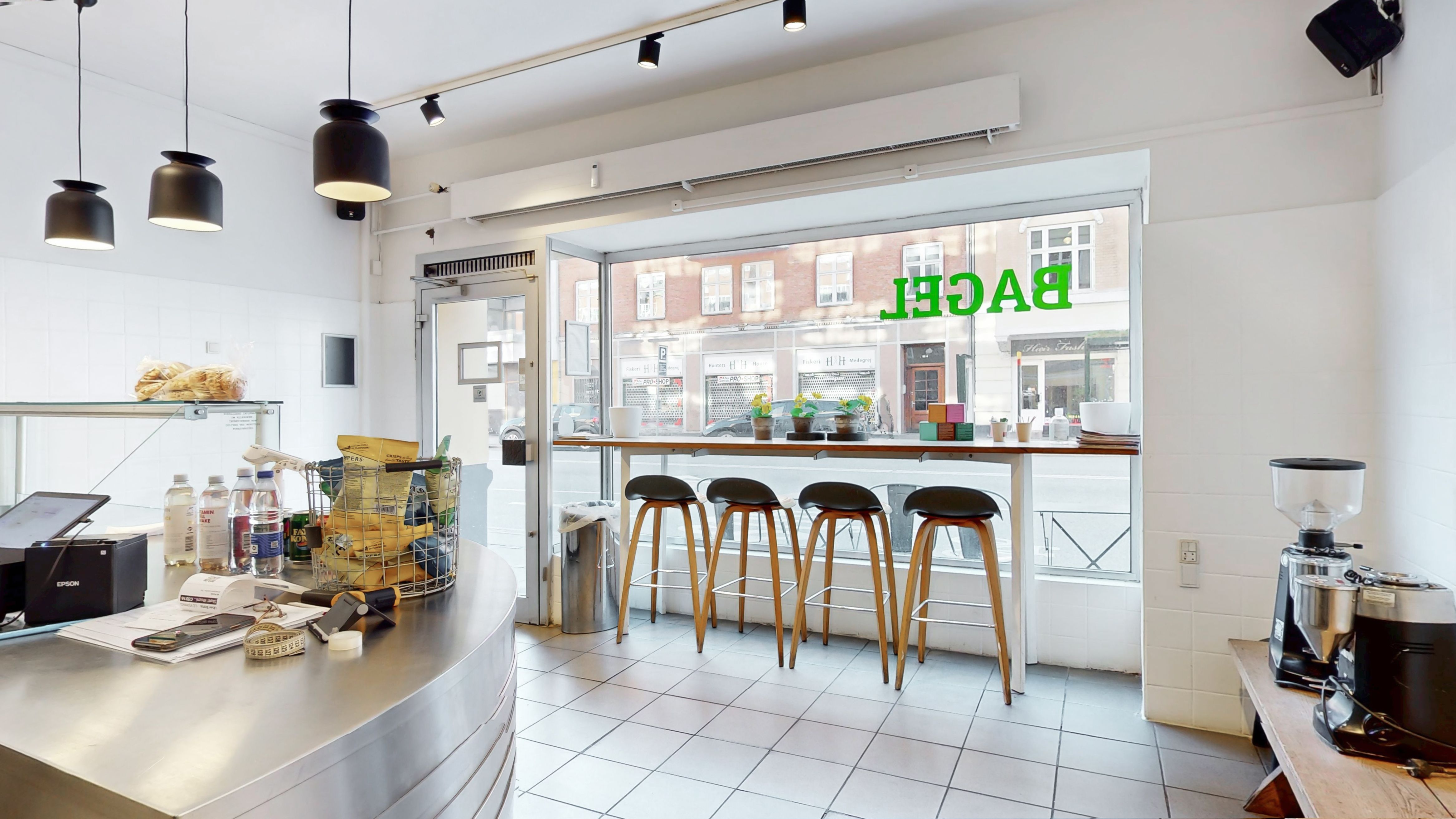 Køkken med bord og stole, og udsigt til gaden med biler og bygninger samt tekst i vinduet og kaffemaskine til højre