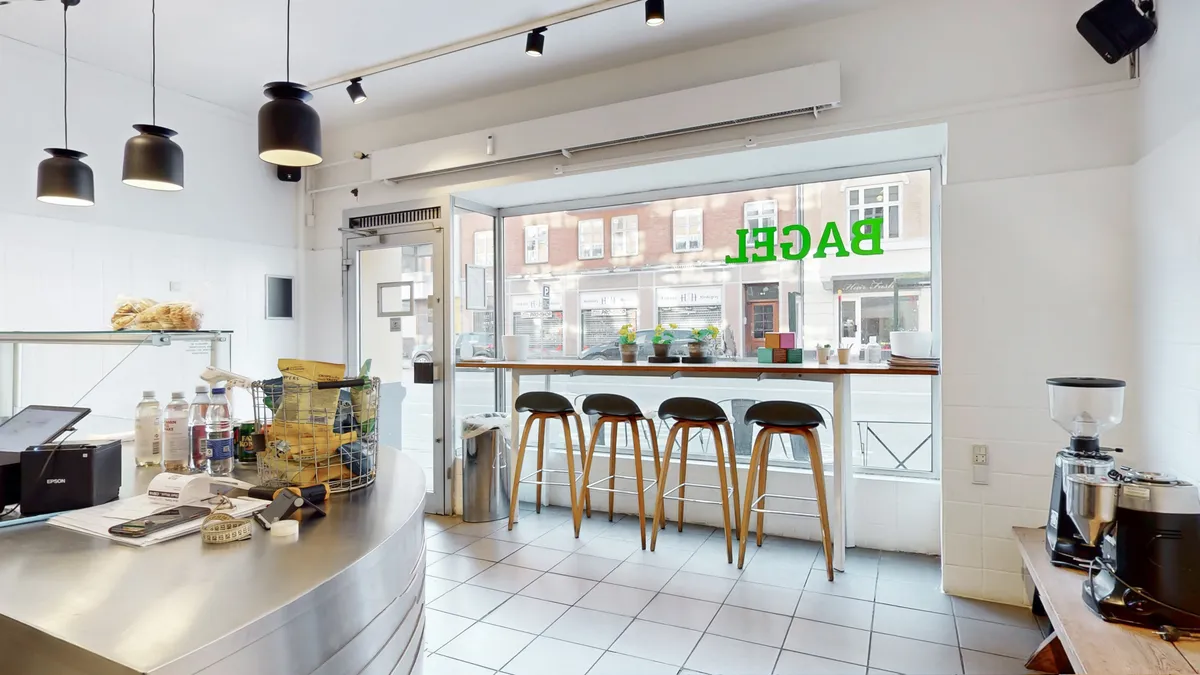 Køkken med bord og stole, og udsigt til gaden med biler og bygninger samt tekst i vinduet og kaffemaskine til højre