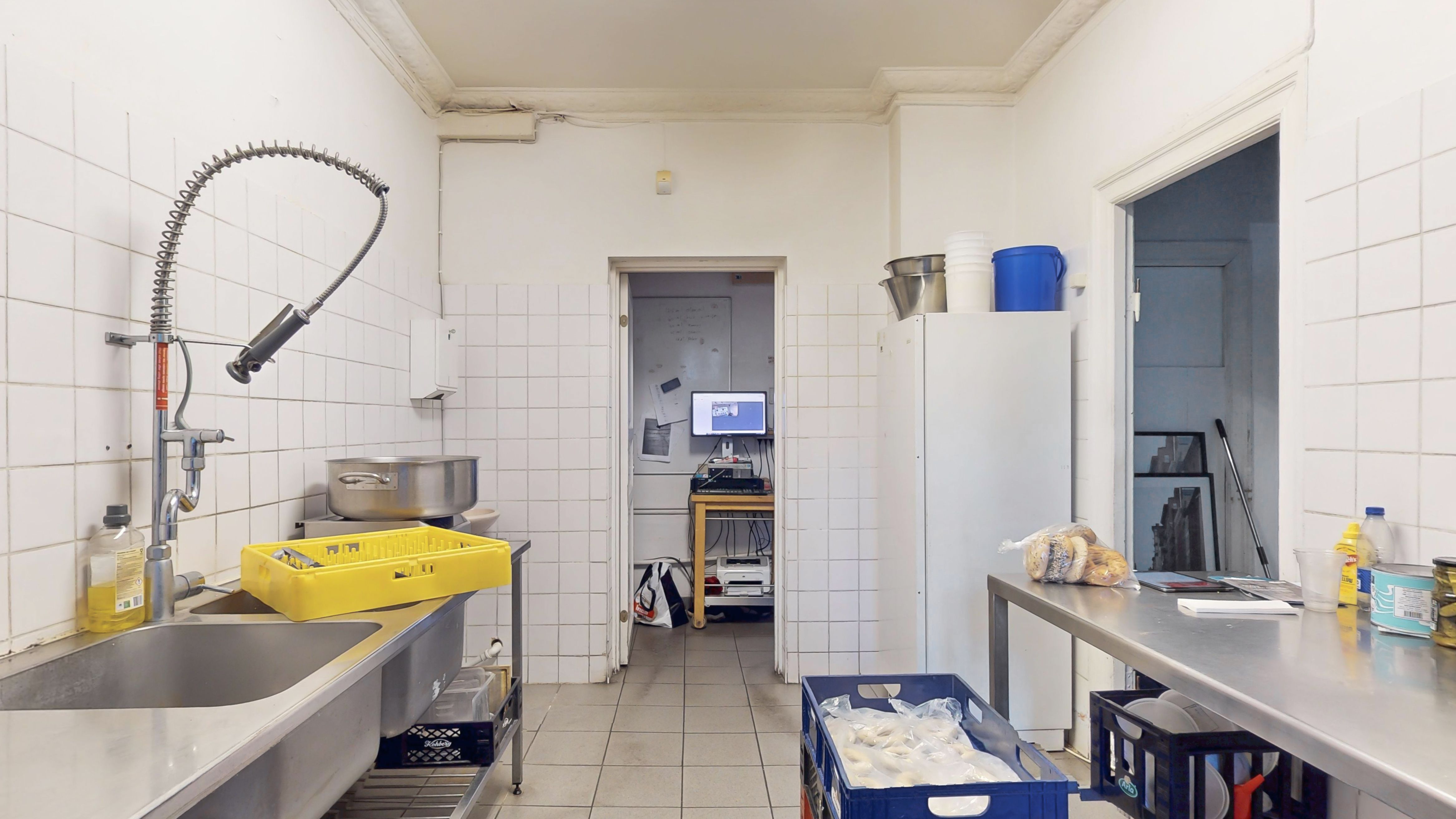 Køkken med to vaskebassiner og vandhane samt en lang metallbord og plastkurve med indpakket mad. Der er også en køleskab og en skærm på skrivebord