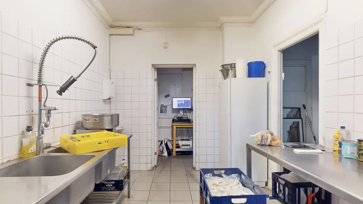 Køkken med to vaskebassiner og vandhane samt en lang metallbord og plastkurve med indpakket mad. Der er også en køleskab og en skærm på skrivebord