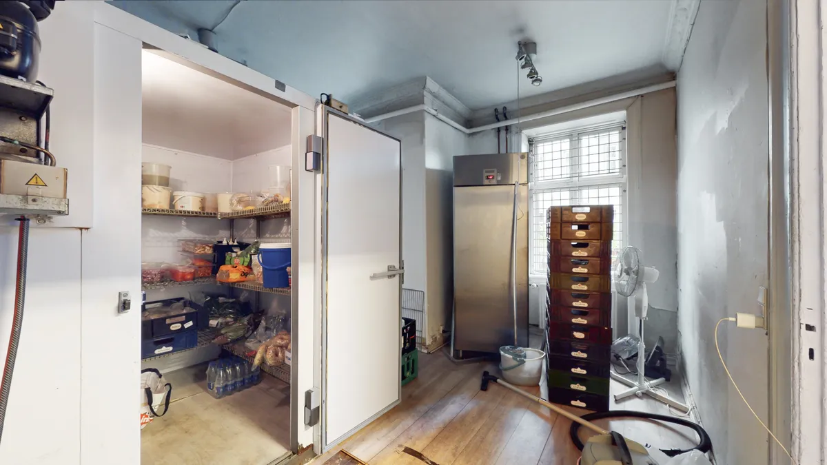 Køkken med udstyr og lager. Køleskab, vinduer og faner samt rækkevis af brædder. Rullebænke med plastflasker og tøjposer.