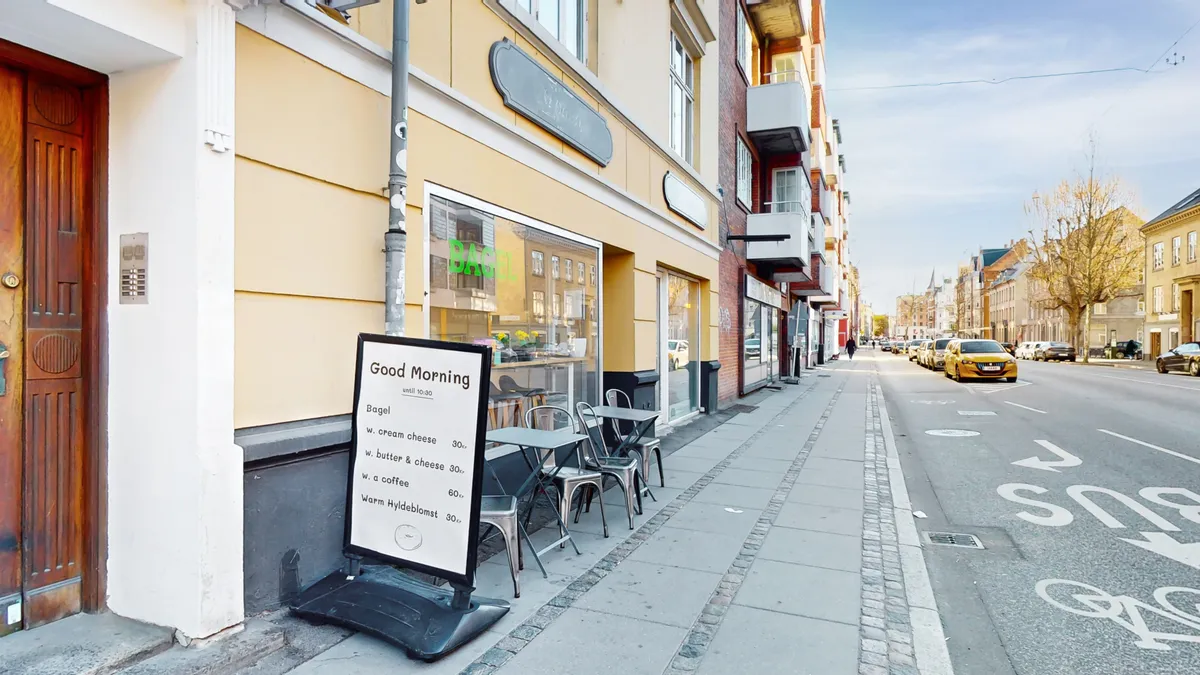 Opholdsområde med træborde og stole uden for et kaffehus, der sælger bøfler og kaffe på glasvinduet. En menutavle med morgenmadsvalg og priser står på fortovet.