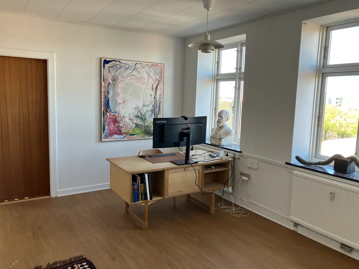 Kontor med skrivebord, monitor og kunstværk samt statue. Smukke vinduer med udsigt til grønne træer og bygninger. Simpel indretning med lyse vægge og trægulv.