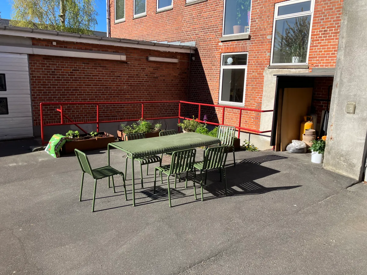 Kontorbord med fire stole og en lang grøn bord placeret i en gårdhave med rød stående skur og en rød metalbøjle