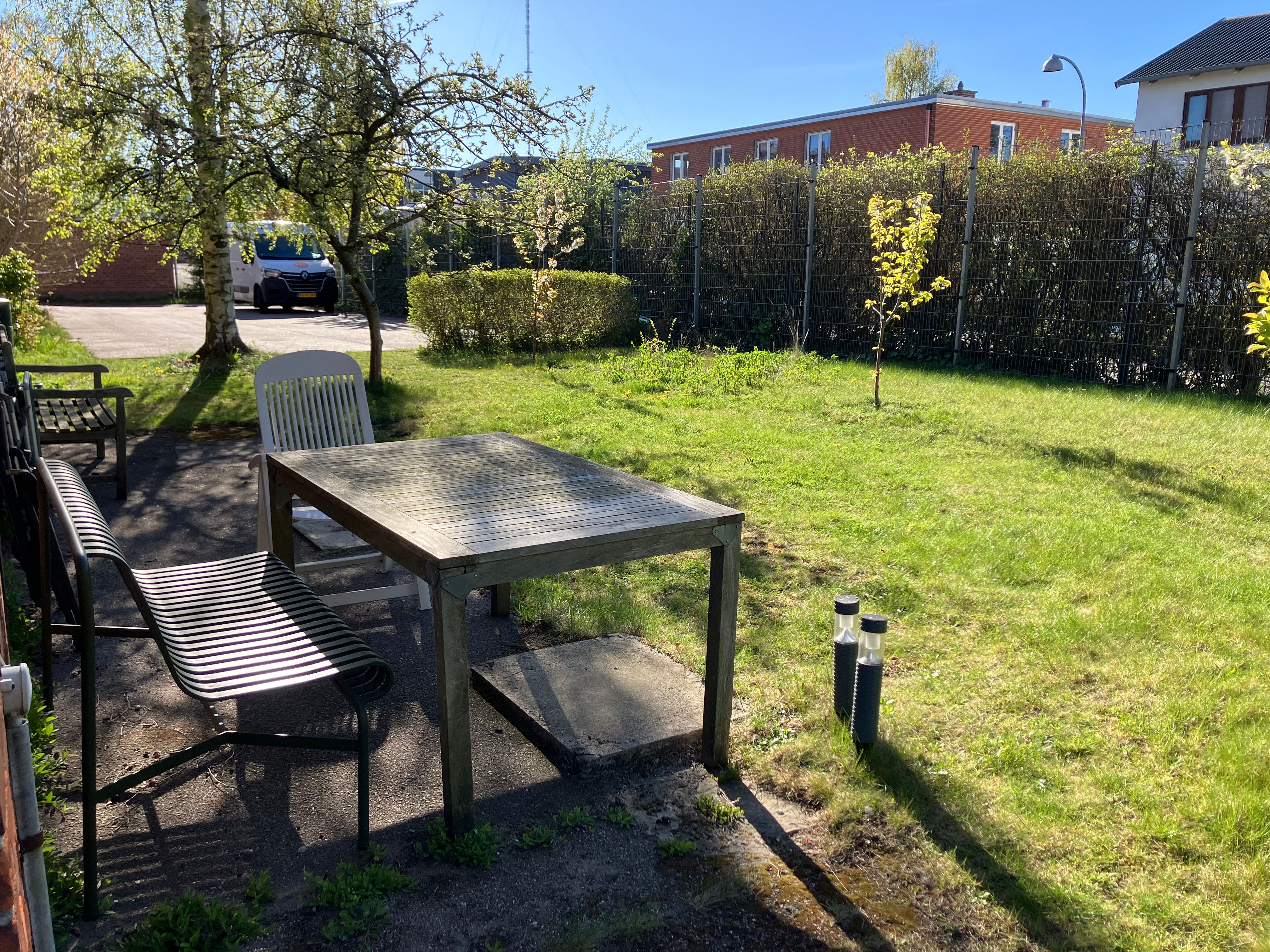 Arbejdsfællesskab med møbelsæt og udendørs parkeringsplads i Søborg. Vandrette rækker af buske og høje røde bygninger i baggrunden.