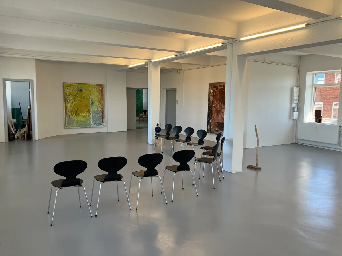 Kunstnerstudie med lyse møbler og lyse loftsbjælker. Værelse med lysegulv og møbler samt store malerier på væggen.