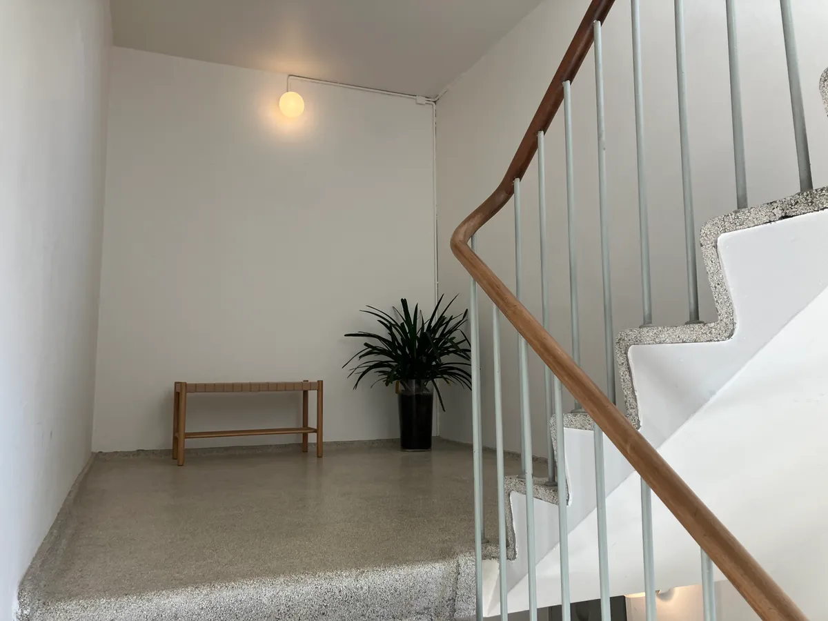 Minimalistisk design med hvid malet væg, lyse gulv og trapper med moderne skrå rækværk og håndtag af metal og træ. Et bord med en plante og en hængende lyspære forstærker minimalistisk følelse.