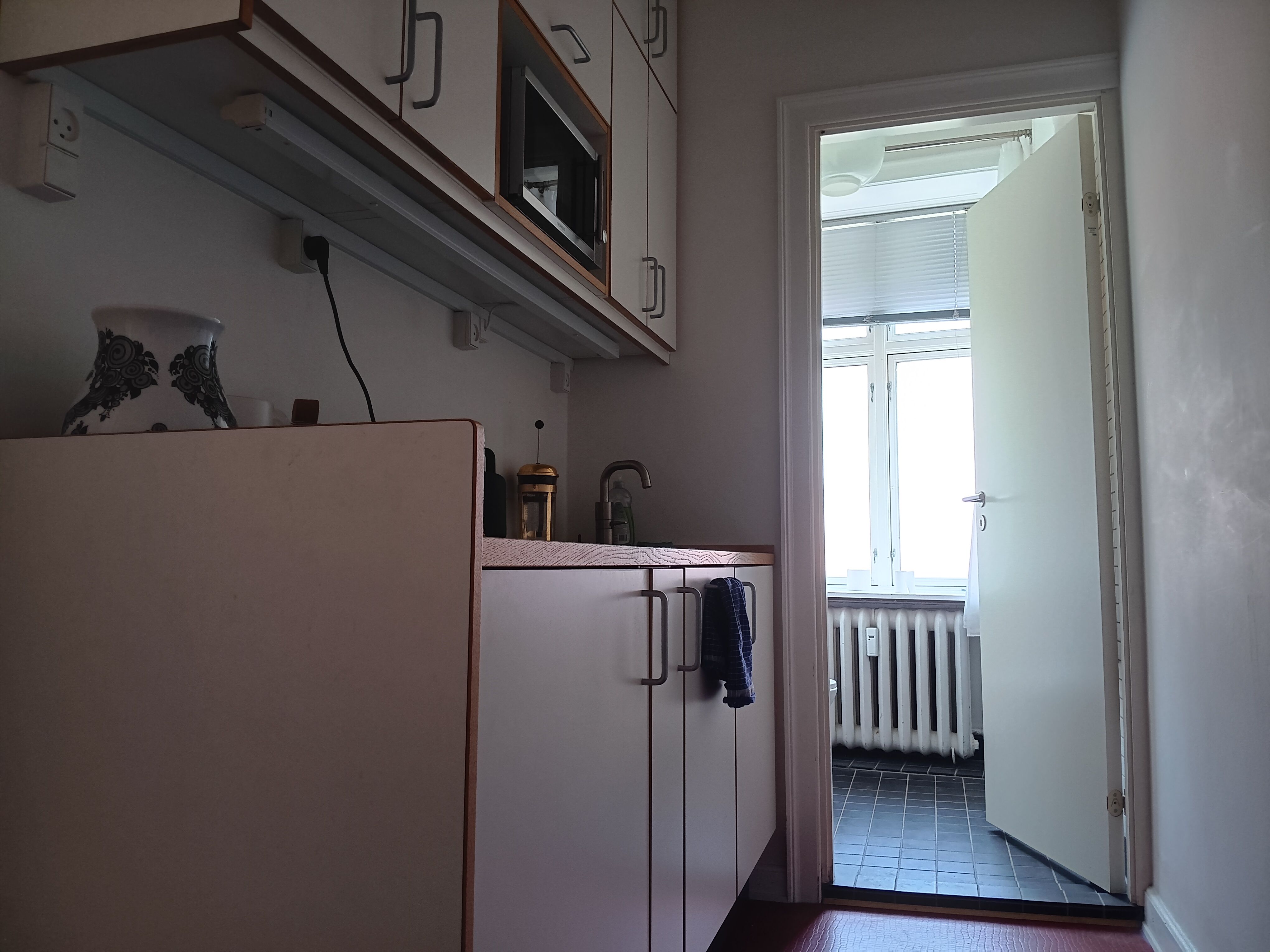 Lyst køkken med højloftede gulv og lyse vinduer. Moderne køkkenudstyr og lyse, hvide vægge. Åbent dør til et andet rum med radiator og lyse vinduer.
