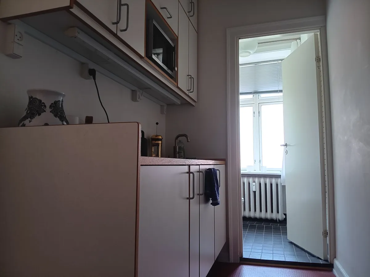 Lyst køkken med højloftede gulv og lyse vinduer. Moderne køkkenudstyr og lyse, hvide vægge. Åbent dør til et andet rum med radiator og lyse vinduer.