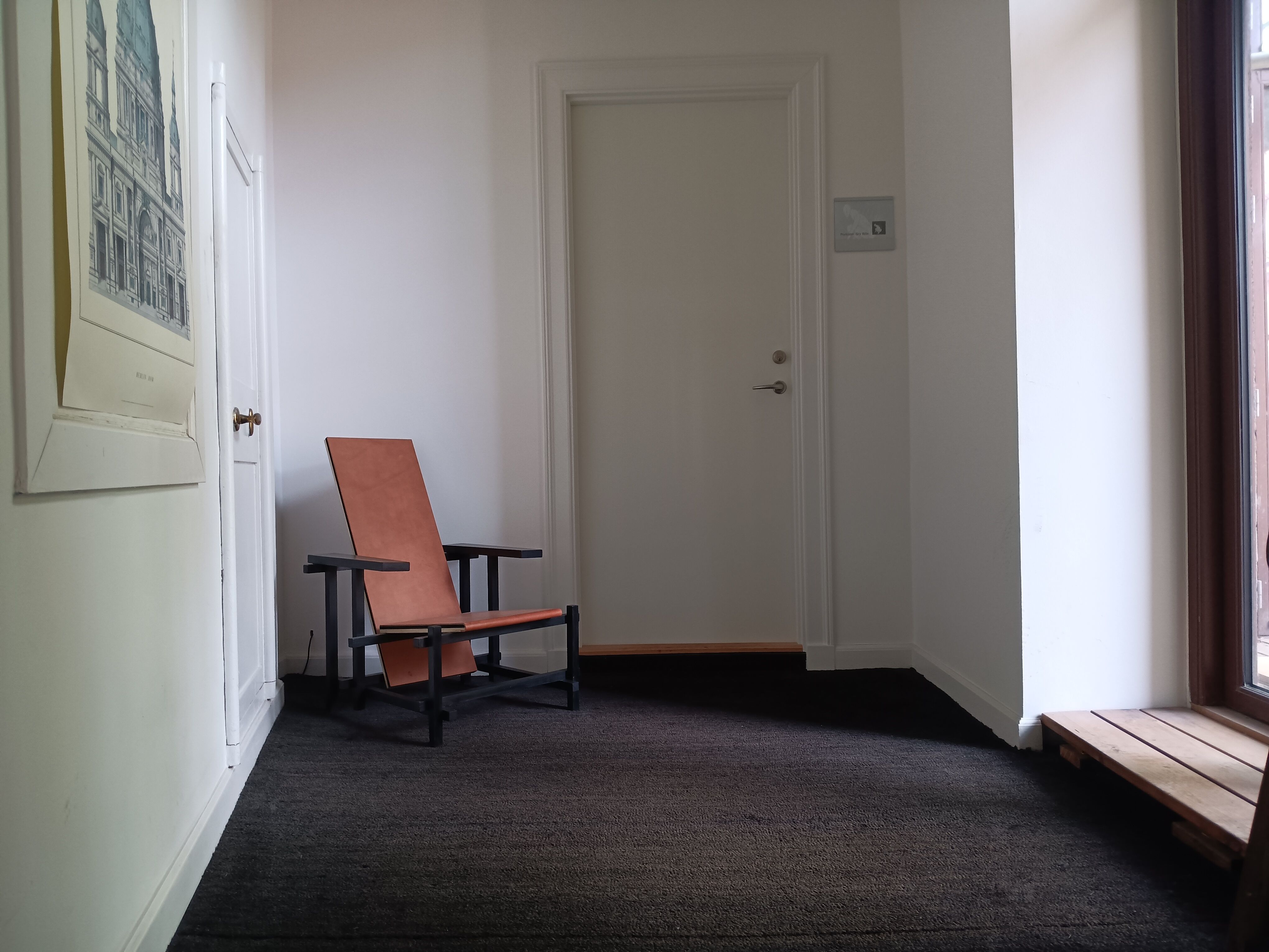 En rummelig og lys hall med trægulv, hvor der står en rusten lænestol og et hængende maleri. Der er et hvidt dørblad og en træbænk ved siden af vinduet.
