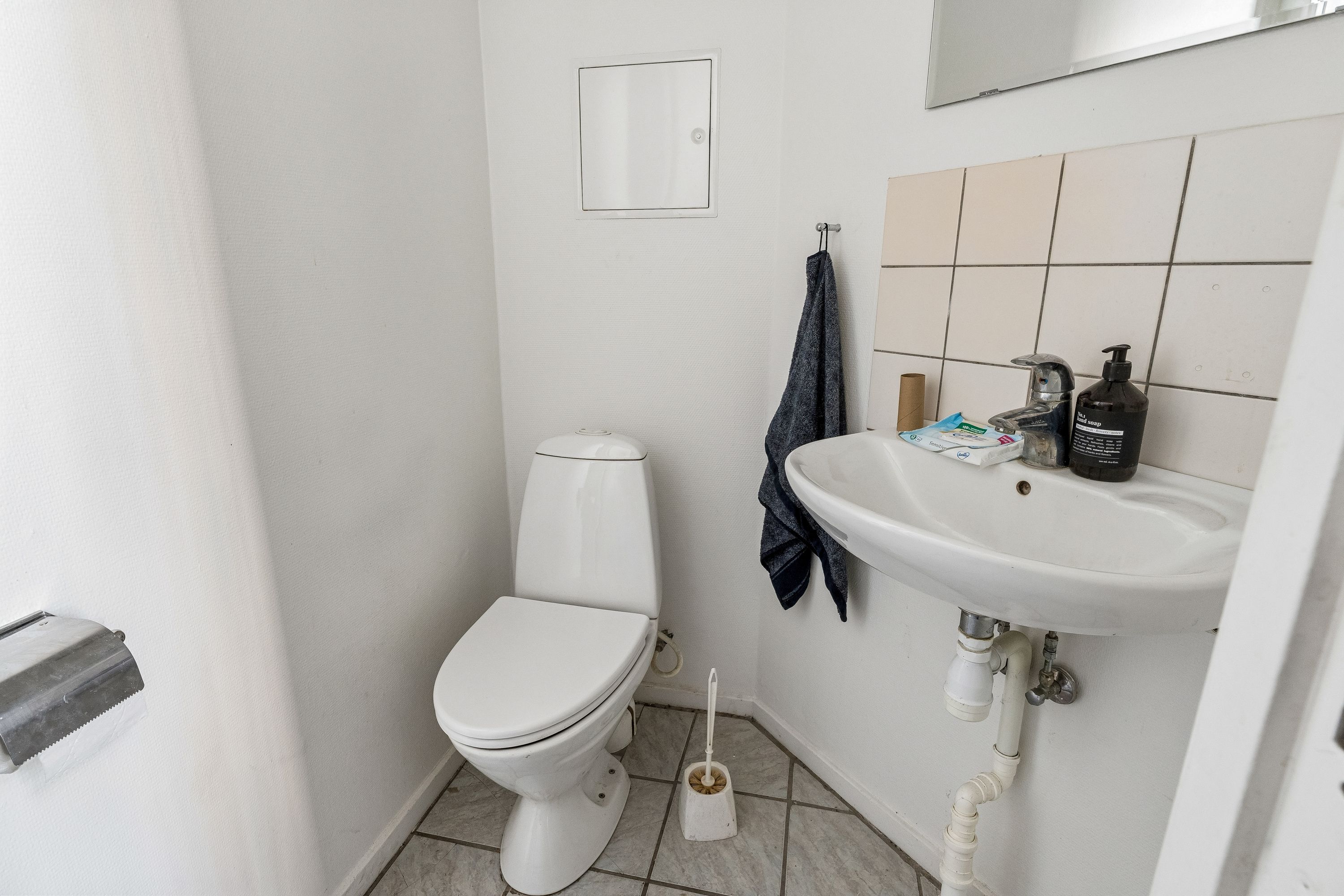 På et lille hvidt badeværelse er der et toilet, håndklæde og et håndvask med sæbe og en børste. På væggen er der en spejl og en toiletpapirholder.