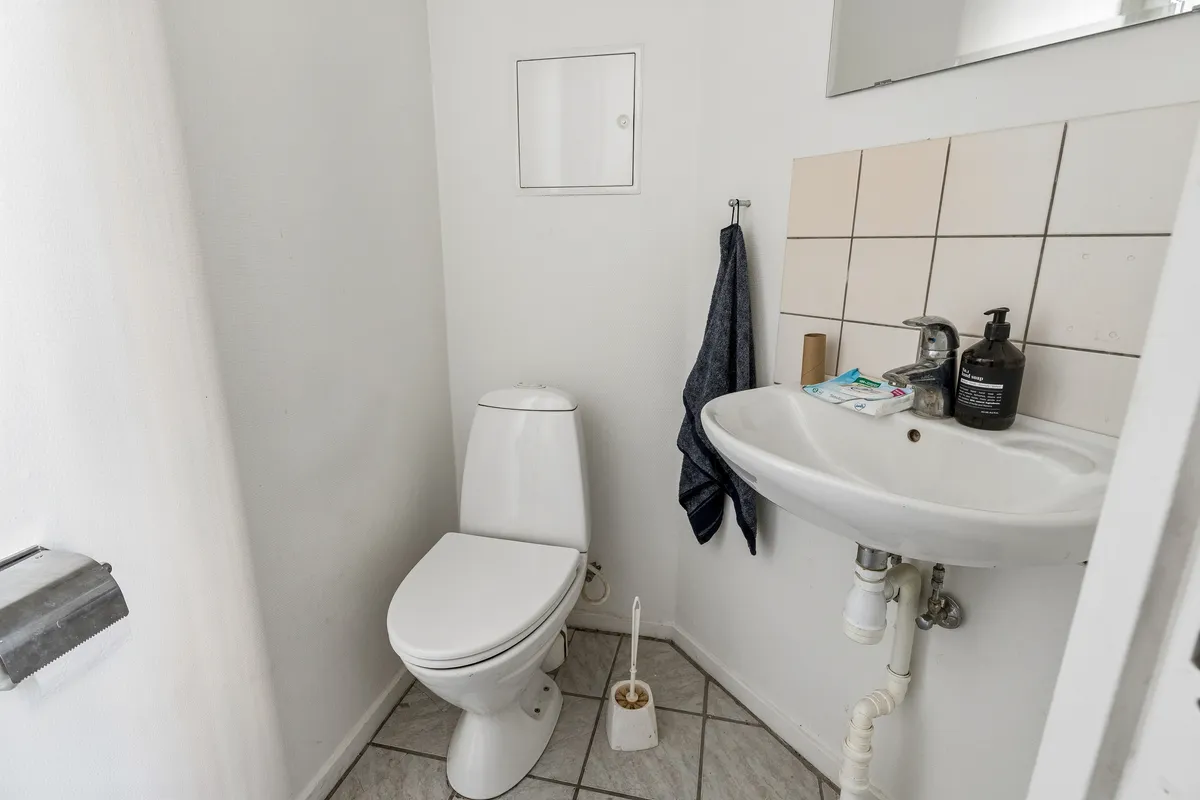 På et lille hvidt badeværelse er der et toilet, håndklæde og et håndvask med sæbe og en børste. På væggen er der en spejl og en toiletpapirholder.