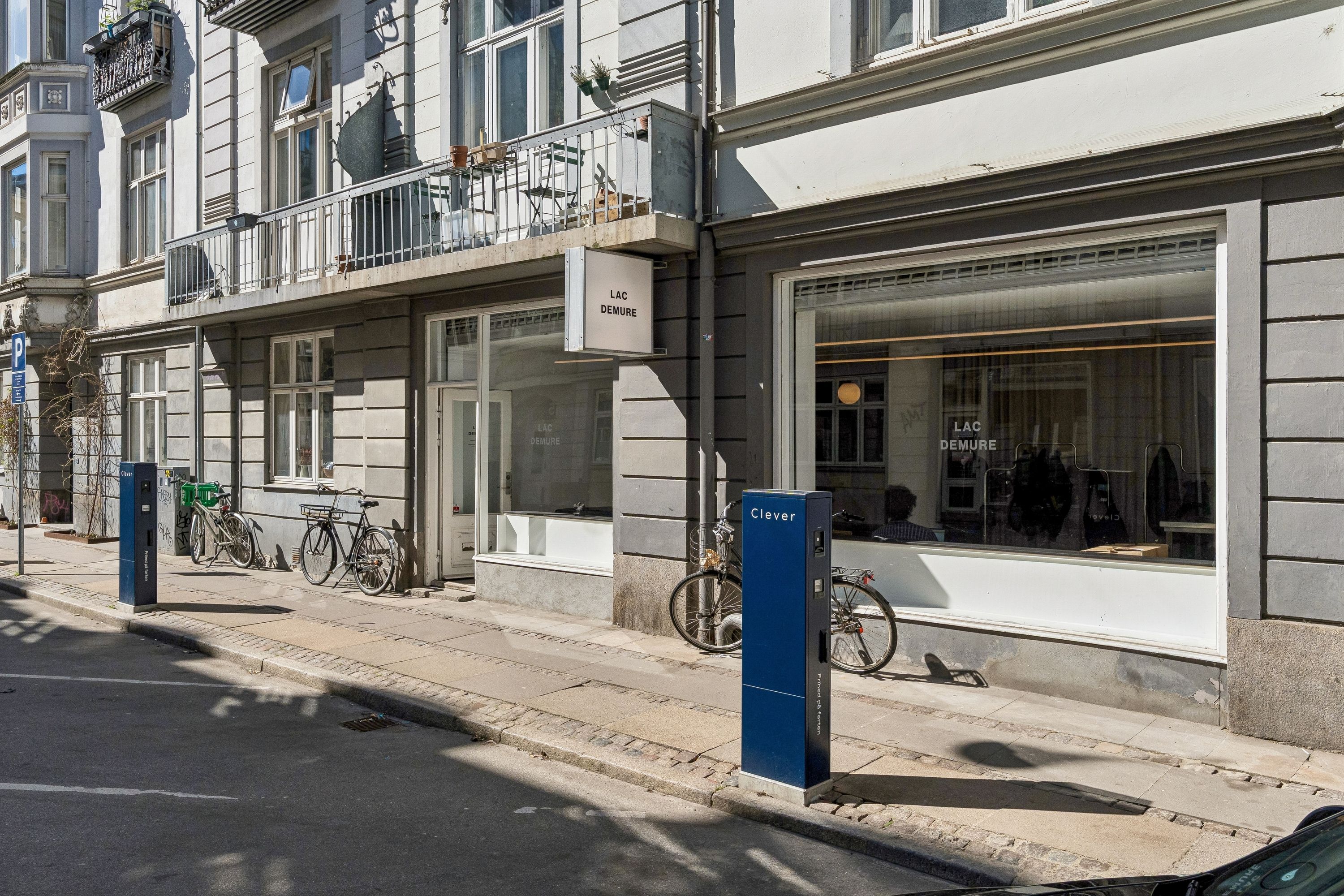 Lejlighed til leje i 4. sal på Westend 4 i 1661 København. Butikker med vinduer og cykler parkeret foran. Bygningen er gulmalet med terrasser og planter.
