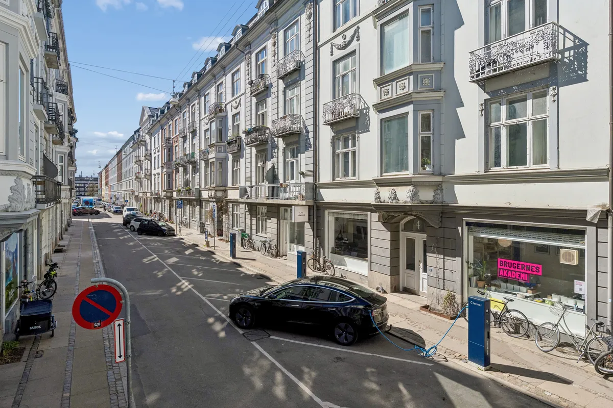 En bil er tilsluttet ladestanderen foran Brugernes Akademi i København. På gågaderne står cykler og biler parkeret, og der er ladestander og snavs på fortovet.