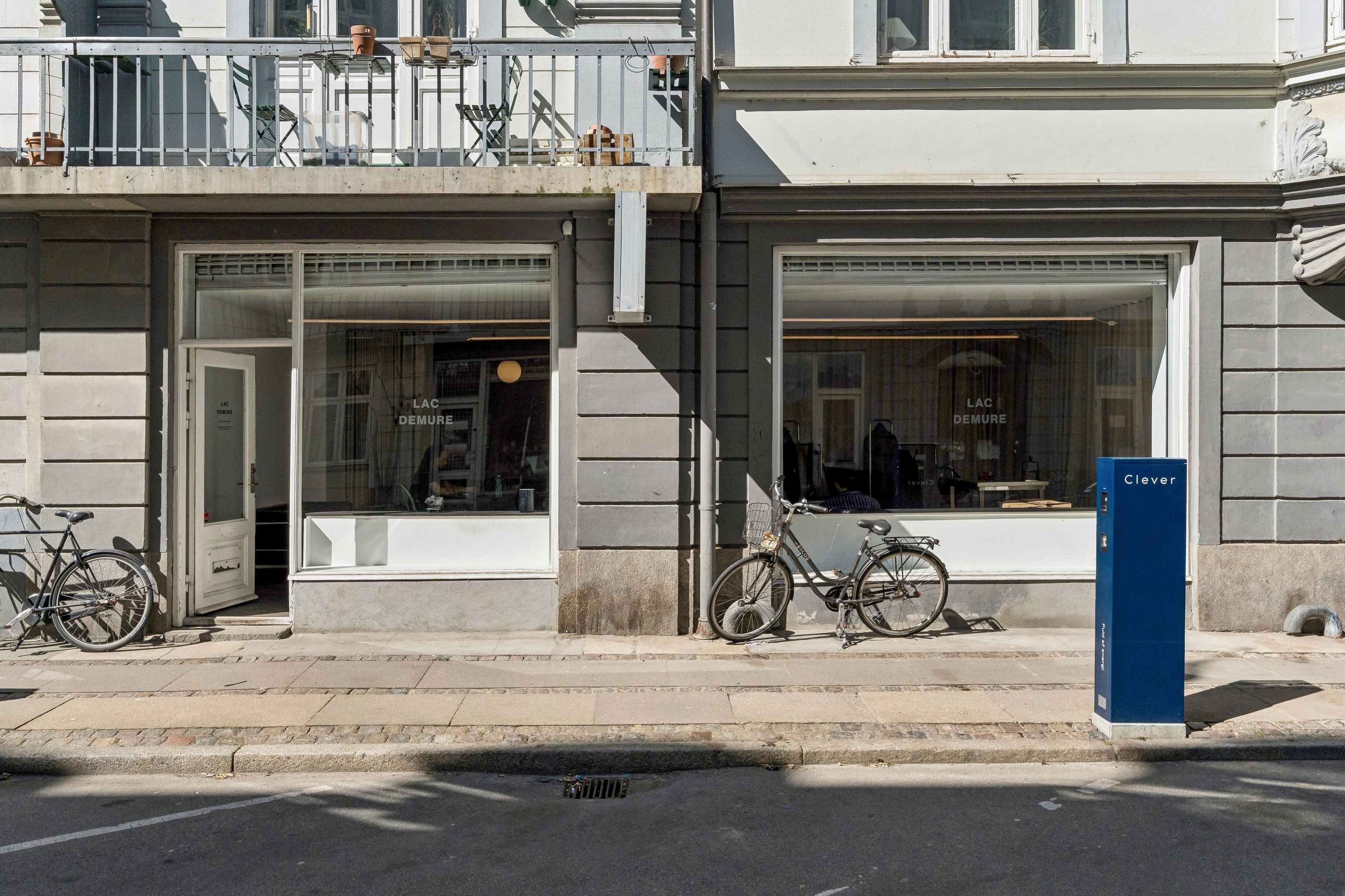 To cykler parkeret foran en glasdør og et vindue med teksten Lac Demure. Der er en Clever kasse foran døren. Der er også en balkon med møbler og potter