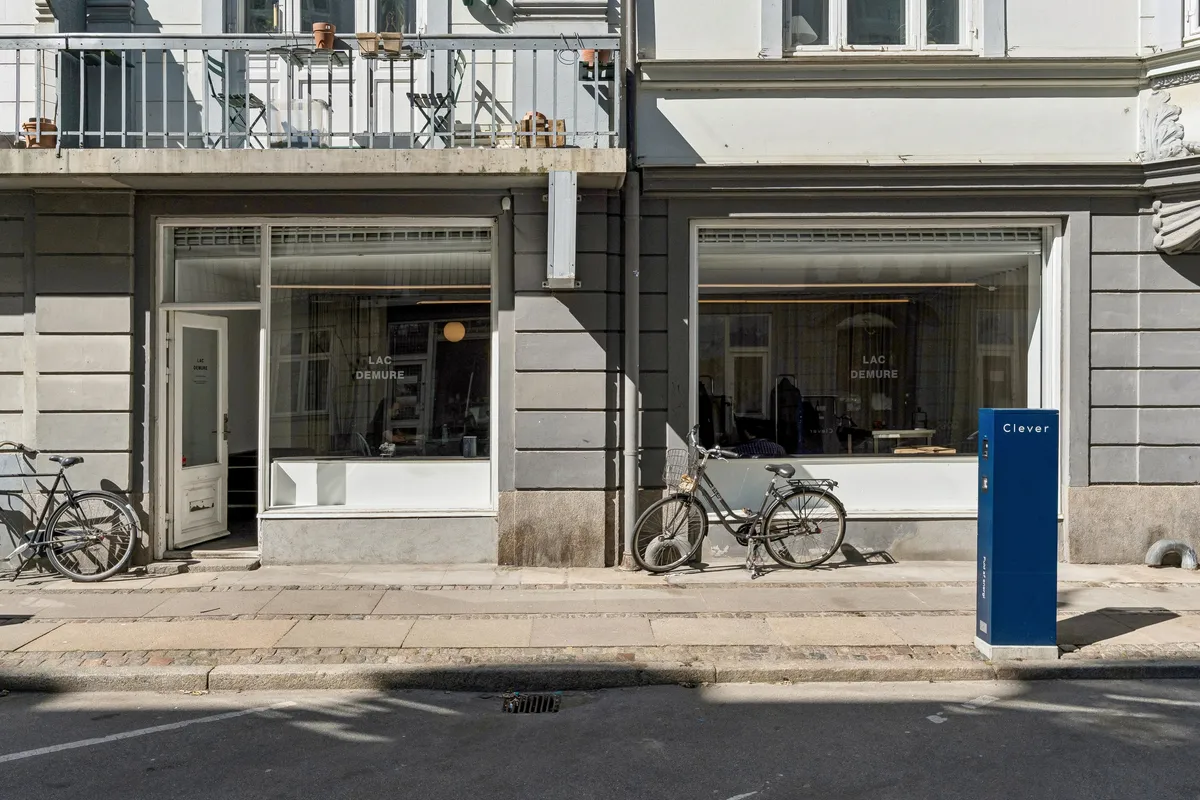 To cykler parkeret foran en glasdør og et vindue med teksten Lac Demure. Der er en Clever kasse foran døren. Der er også en balkon med møbler og potter