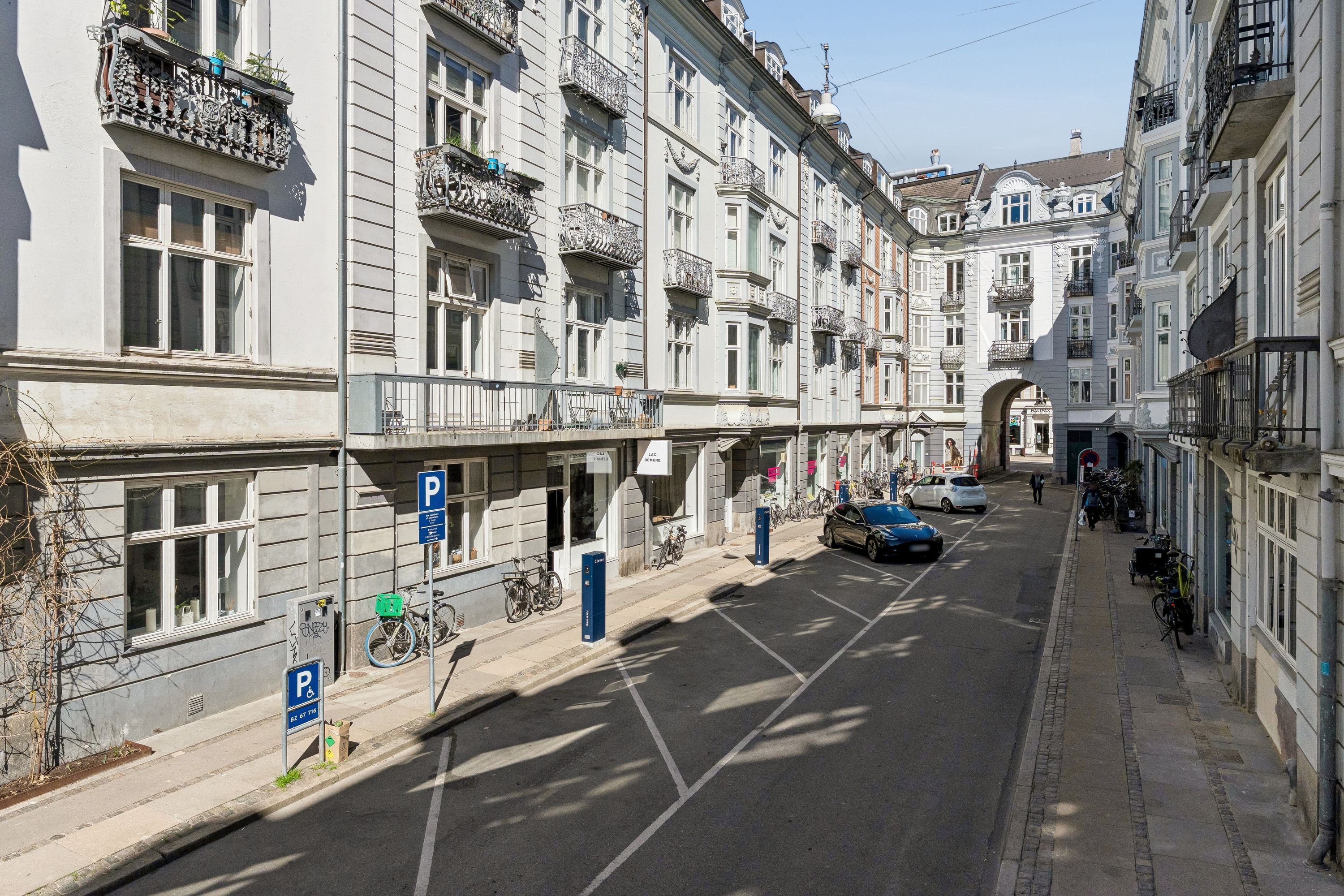 En bilparkeringsplads med parkerede biler og cykler langs gaden, hvor der er parkerede biler og folk går på fortovet. Der er to parkeringsskilte, et hvidt og et blåt