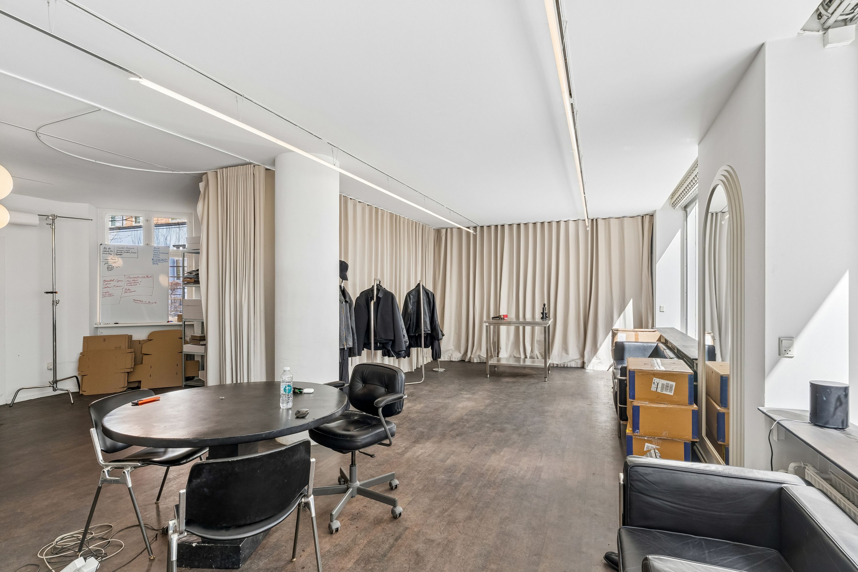Butikslejemål med 89 m² rum, højloft og store vinduer. Rundt borde, læderstole og hængeklæder. Værelse med skrivebord, højtalere og lysekroner.
