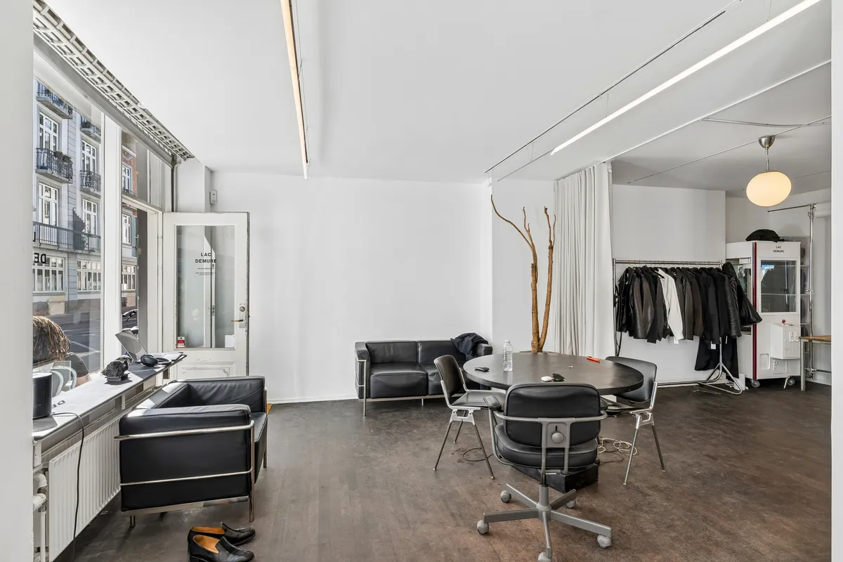 Butik med fuldt glasvindue og moderne møbler og træelementer. Sort sofa, bord og stole samt hængeklæder og tøjskabe. Lampe på loftet.