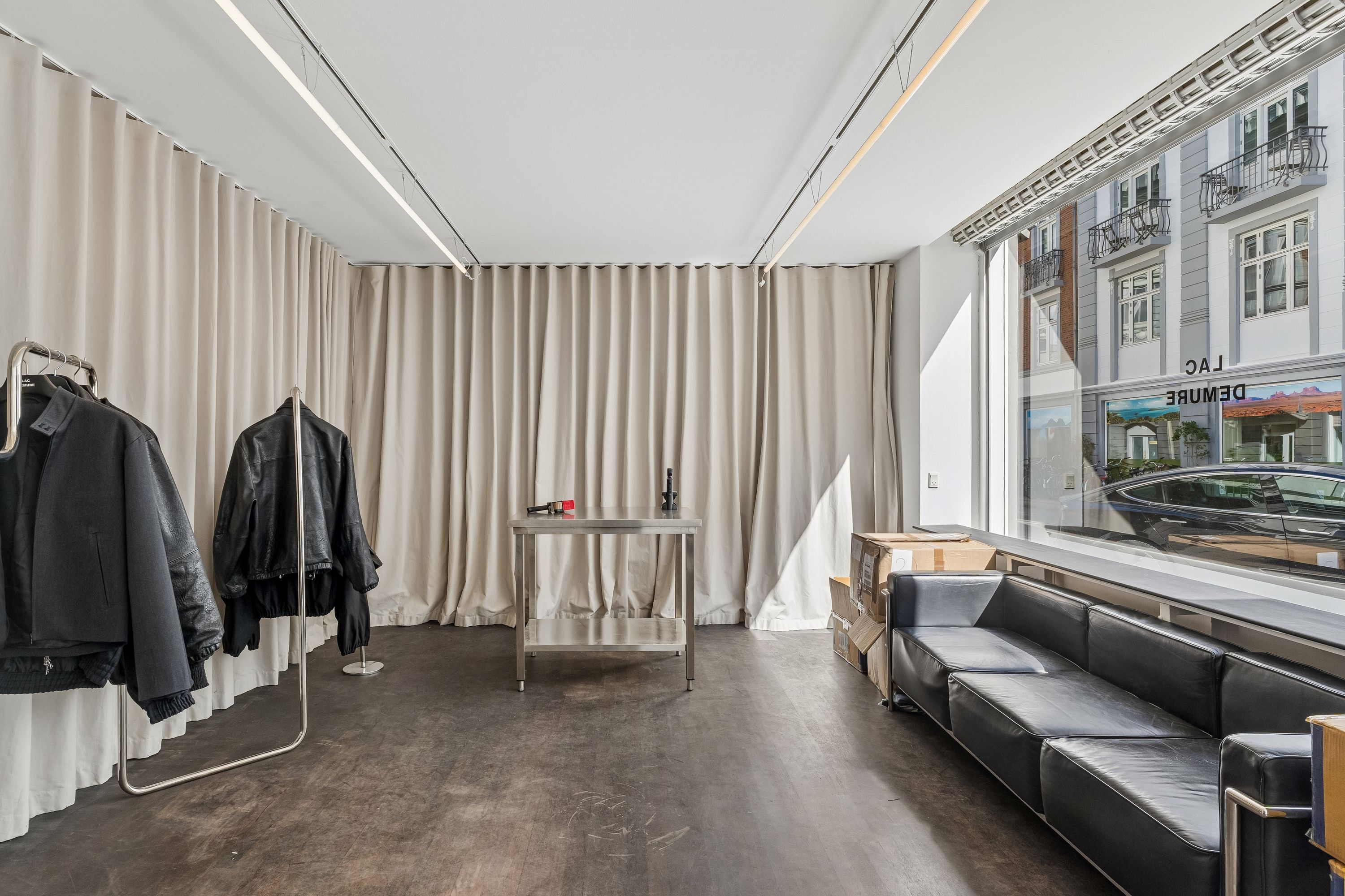 En moderne butikslokale med store hvide gardiner og trægulv, udstyret med en lædersofa og en stålbord, og udsigt til en parkeret bil og bygninger med balkoner